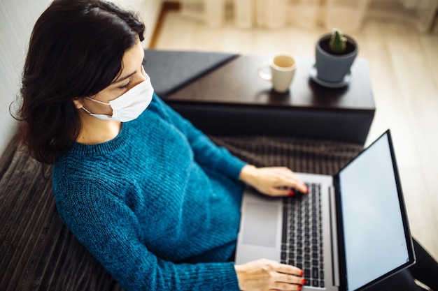 Businesswoman stays home and works during coronavirus epidemia quarantine. Female worker wearing a medical mask and typing on a laptop. Covid-19 pandemia spread prevention concept.