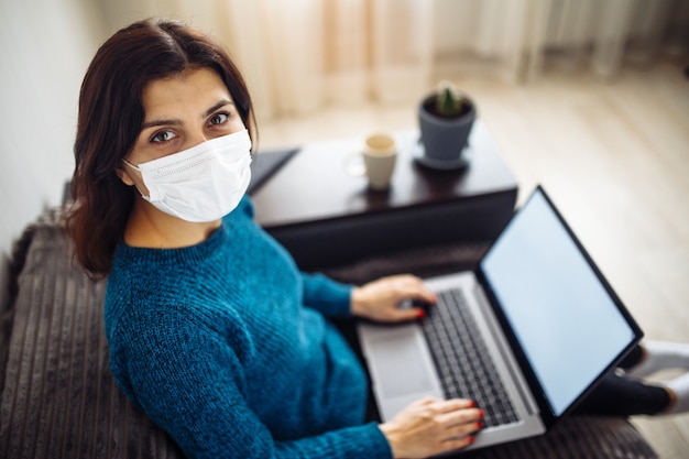 Businesswoman stays home and works during coronavirus epidemia quarantine. Female worker wearing a medical mask and typing on a laptop. Covid-19 pandemia spread prevention concept.