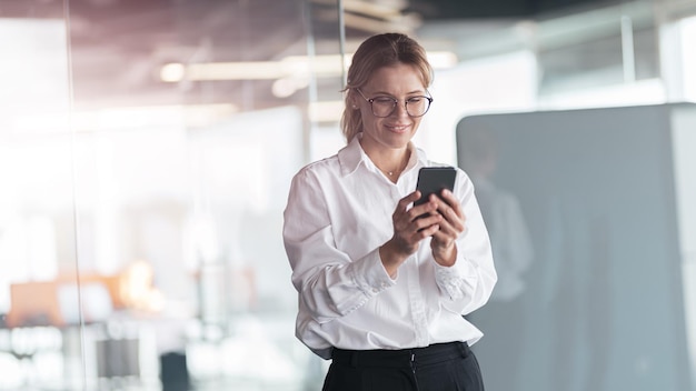 Businesswoman wearing glasses with mobile phone in hand standing in modern office