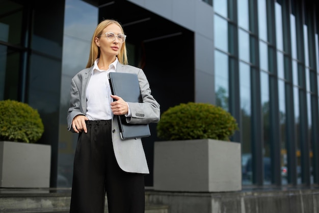 Businesswoman wearing stylish eyeglasses holding laptop looking away standing on the street