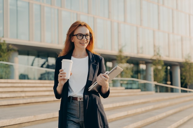 Businesswoman wears elegant formal clothes drinks takeaway coffee carries notebook and modern gadget