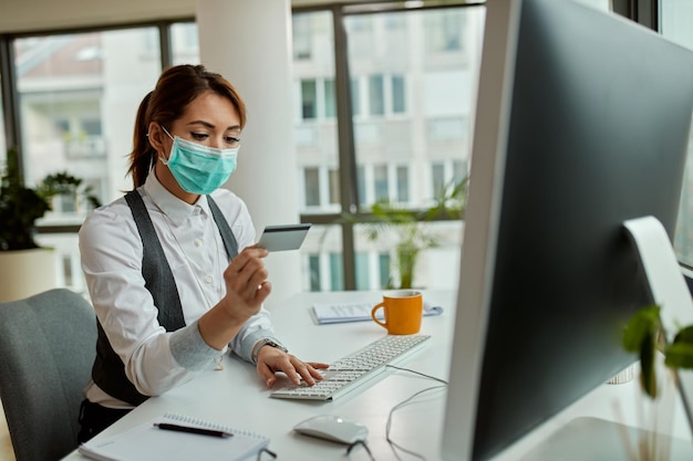 Businesswoman with face mask using credit card while checking her online bank account on a computer in the office