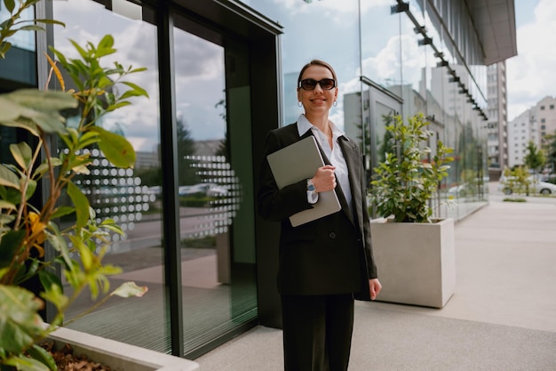 Businesswoman with laptop standing outside modern office building looking confident