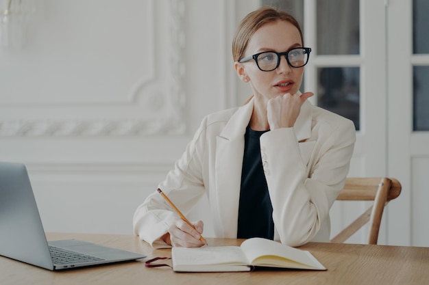 Businesswoman writes business ideas in notebook makes to do list planning day sitting at laptop