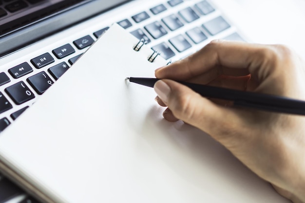 Businesswoman writes with a pen in diary on laptop keyboard in a sunny office business and education concept Close up