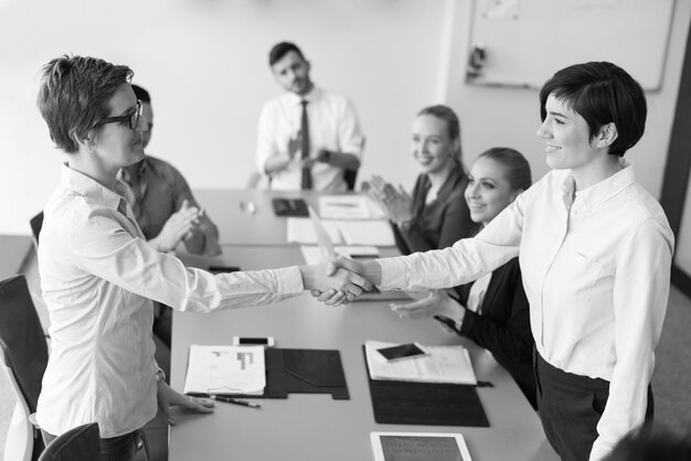 businesswomans handshake on team meeting  with group of people blured in background at modern startup business office interior