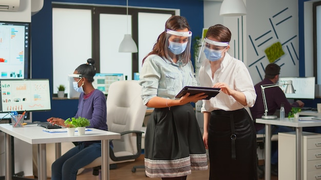 Businesswomen with protection masks against coronavirus standing in workplace talking about company financial datas holding digital tablet. Multiethnic business team working respecting social distance