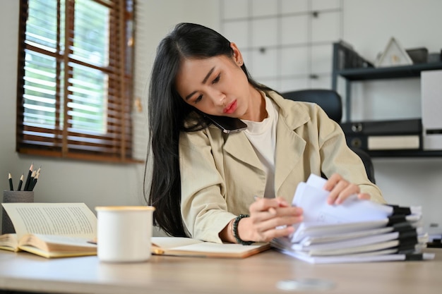 Busy Asian businesswoman on the phone while working on her documents at desk