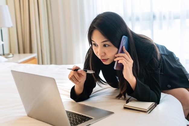 Busy Asian businesswoman working on laptop computer while talking on phone in bed at hotel Business trip people and technology concept copy space