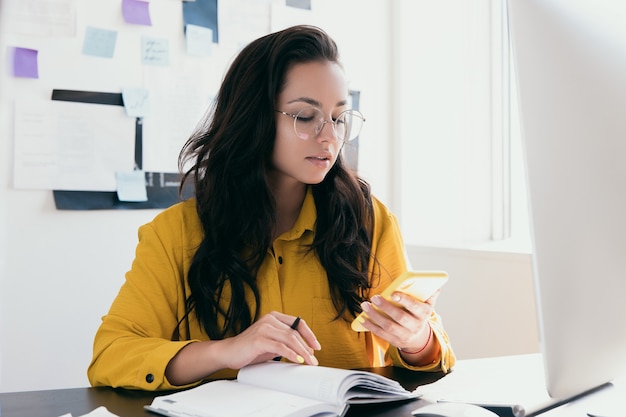 Busy attractive woman in round glasses searching information in web or answered to client by their smatphone