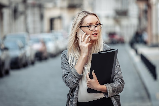 Busy business woman in business suit talking on the phone and holding folder on city street