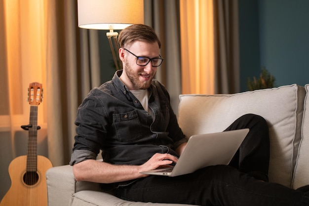 Busy college student with stubble and glasses sits with a smile in the evening on the living room co