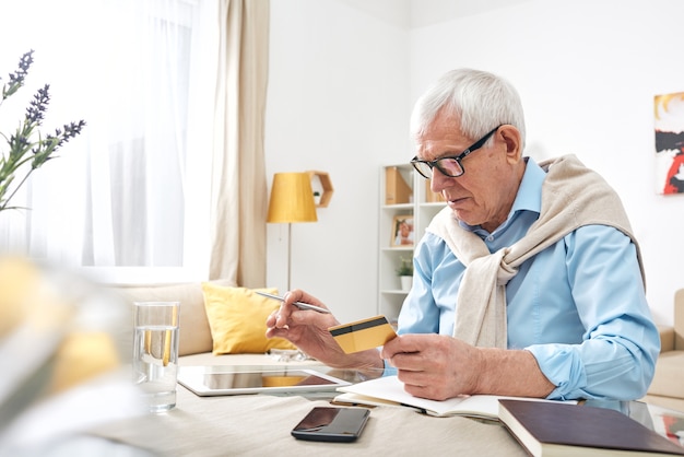 Busy elderly wrinkled man in eyeglasses using digital tablet while checking balance of debit card at home