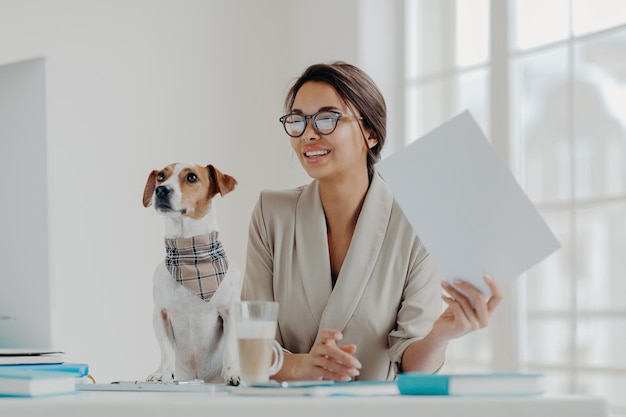 Busy female entrepreneur works with papers prepares business report concentrated in monitor of computer dressed formally dog sits near pose at desktop with notepads around Pet helps working