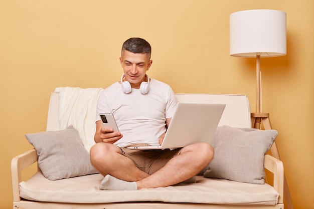 Busy handsome man wearing white Tshirt and shorts sitting on sofa against beige background working on laptop at home using mobile phone while having break