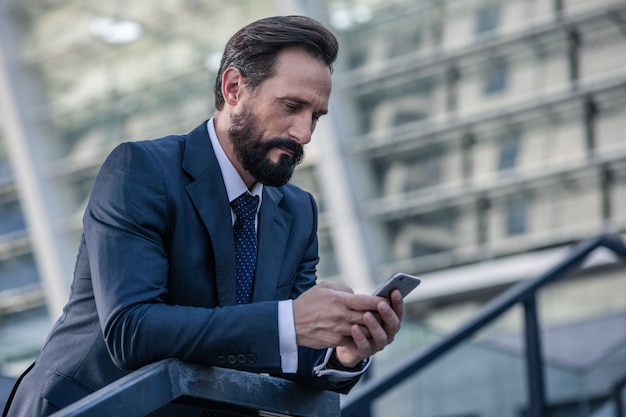 Busy life. Waist up of a bearded concentrated businessman holding a phone while leaning on the handrail