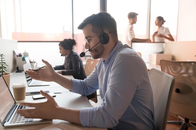 Busy male call center agent in glasses wearing headset talk use laptop consulting client online concentrated man telemarketer in earphones speak helping customer working on computer in office