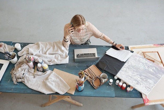 Photo busy mature woman in eyeglasses sitting at desk and examining papers while talking to customer by phone in art studio