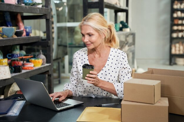 Busy mature woman, small business owner smiling and drinking coffee while working on the laptop 