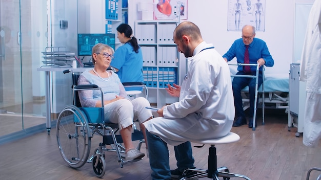 In busy modern private recovery clinic or hospital doctor is talking with disabled patient in wheelchair while nurse is bringing a man with walking disabilities in. Health care medicine support assist