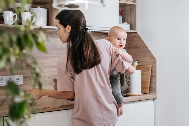 Busy mother cleaning kitchen with baby on her arms. Woman cooking and holding infant, tired woman doing daily homework with kid.