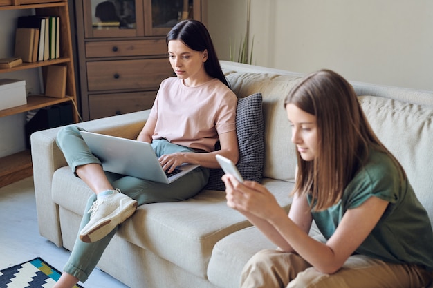 Busy mother sitting on sofa and checking email on laptop while her daughter surfing net on smartphone