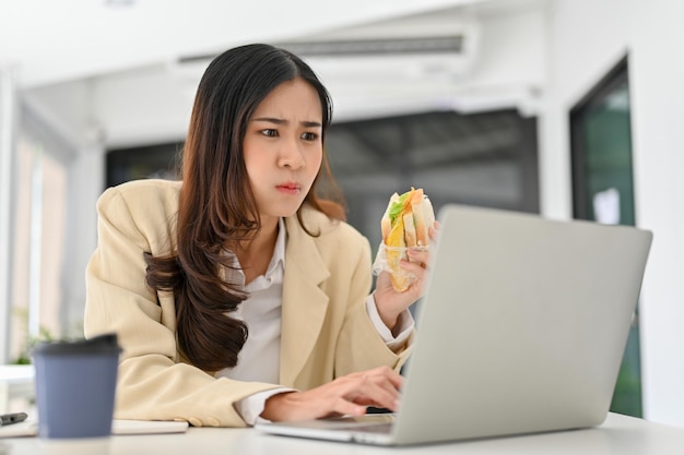 Busy and overworked Asian businesswoman eating a sandwich while using laptop