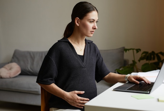 Busy pregnant young Caucasian woman sitting at table in living room and using laptop while working remotely