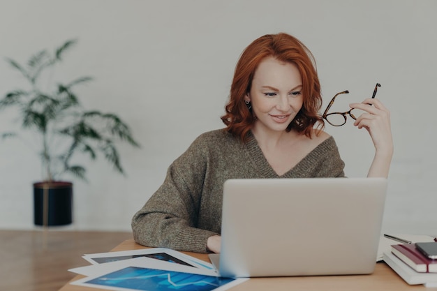 Busy redhead freelancer clad in a warm sweater works on laptop analyzing data and preparing projects amidst paper documents utilizing the internet