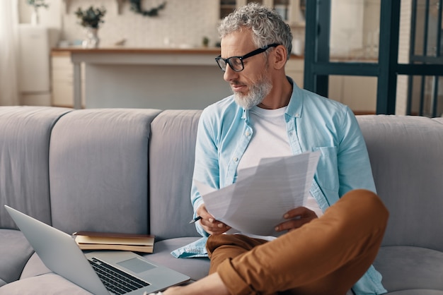 Busy senior man checking the papers and using laptop while sitting on the sofa at home