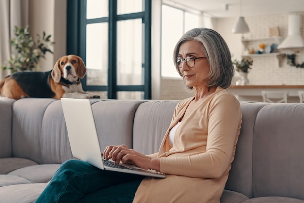 Busy senior woman in casual clothing using laptop and smiling while sitting on the sofa at home