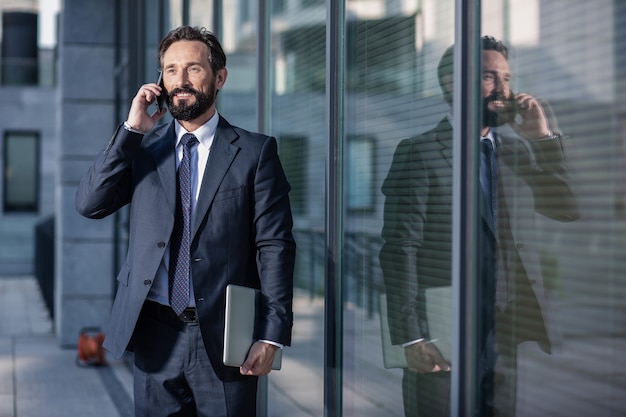 Busy worker. Pleasant adult office worker talking on phone while holding a laptop