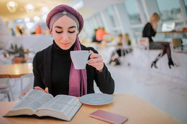 Busy young arabain woman read book and drink coffee. She it turning picture. She sits in side. Woman is busy