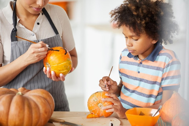 Busy young black woman and her curlyhaired son standing at counter and painting pumpkins