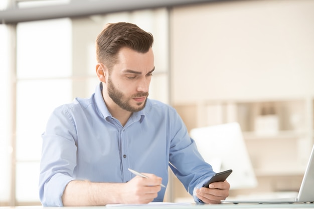 Busy young employee scrolling or messaging in smartphone while planning work by desk in office