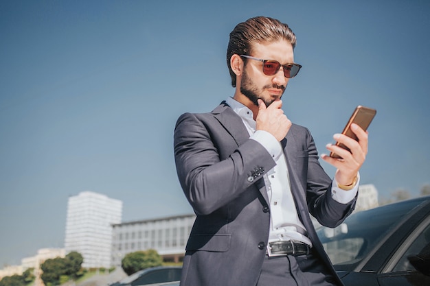 Busy young man looks at phone he holds in hands. Guy has another hand on chin. He thinks. Young man stands at black car.