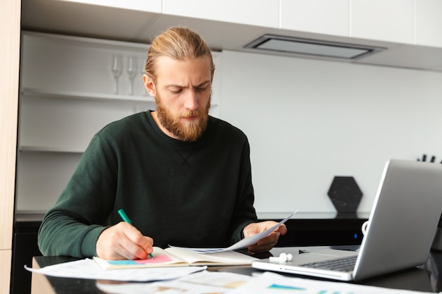 Busy young man working on laptop computer at home, sitting at the kitchen