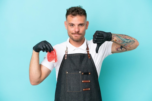 Butcher caucasian man wearing an apron and serving fresh cut meat isolated on blue background showing thumb down with negative expression