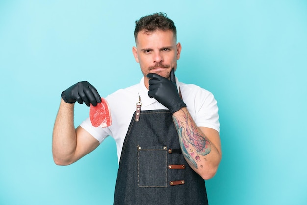 Butcher caucasian man wearing an apron and serving fresh cut meat isolated on blue background thinking