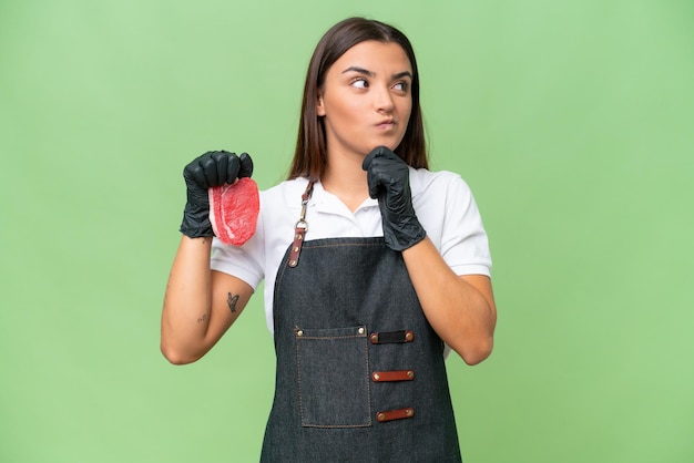 Butcher woman wearing an apron and serving fresh cut meat isolated on green chroma background having doubts and thinking