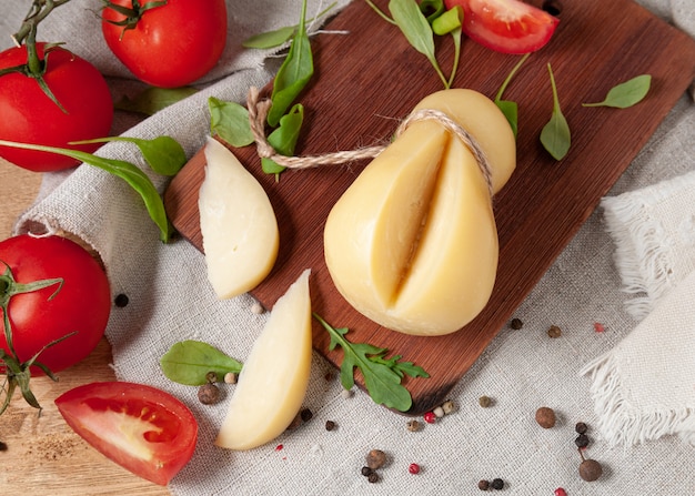 Caciocavallo cheese lies on a rectangular wooden board. Two pieces are cut off. Decorated with arugula, spices and tomatoes. Background gray linen fabric. View from above.