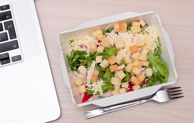 Caesar salad with chicken in takeout packaging with fork and laptop on office table Food delivery and takeout food concept