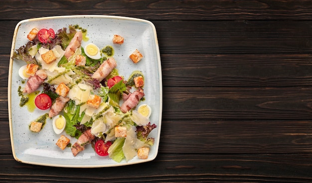 Caesar salad with herbs chicken bacon eggs and tomatoes on a square light plate on a dark background