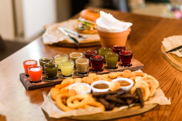 In a cafe with a homely atmosphere a set of alcohol tinctures and a wooden plate with onion rings cheese sticks and balls and sauces are on the table Horizontal photo