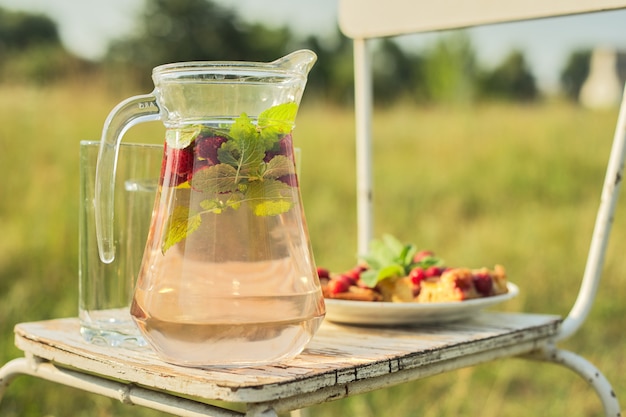 Cake with berries and jug with strawberry mint drink in summer sunny meadow