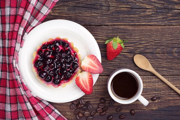 Cake with black currants, strawberry and coffee cup on dark wooden table, top view