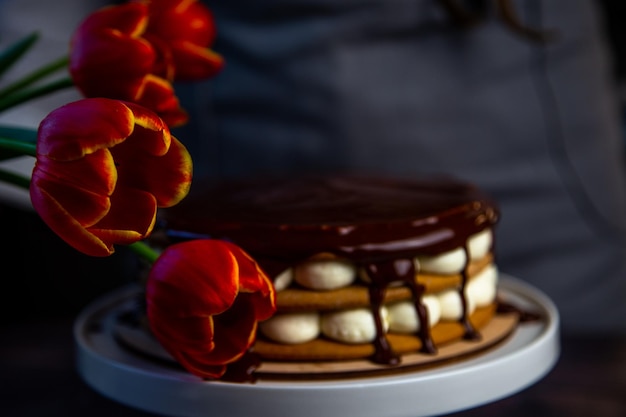 A cake with honey cakes and white cream stands on a white stand and is poured with chocolate on top ...