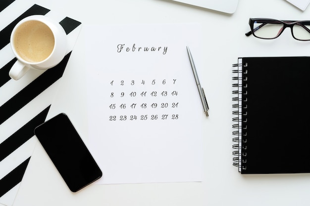 Calendar of February on white desktop flat lay with a cup of coffee and a notebook, work space