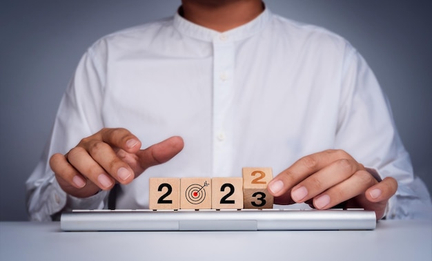 The calendar year 2022 changes to new year 2023 business goal and success plan concept Wooden blocks turn by hand for the transition from 2022 to 2023 with target icon on keyboard computer on desk