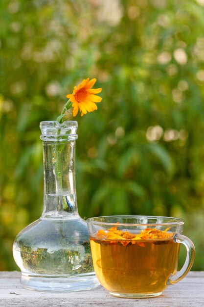 Photo calendula flower with a stem in a glass flask and same flowers beside, cup of tea on wooden boards.